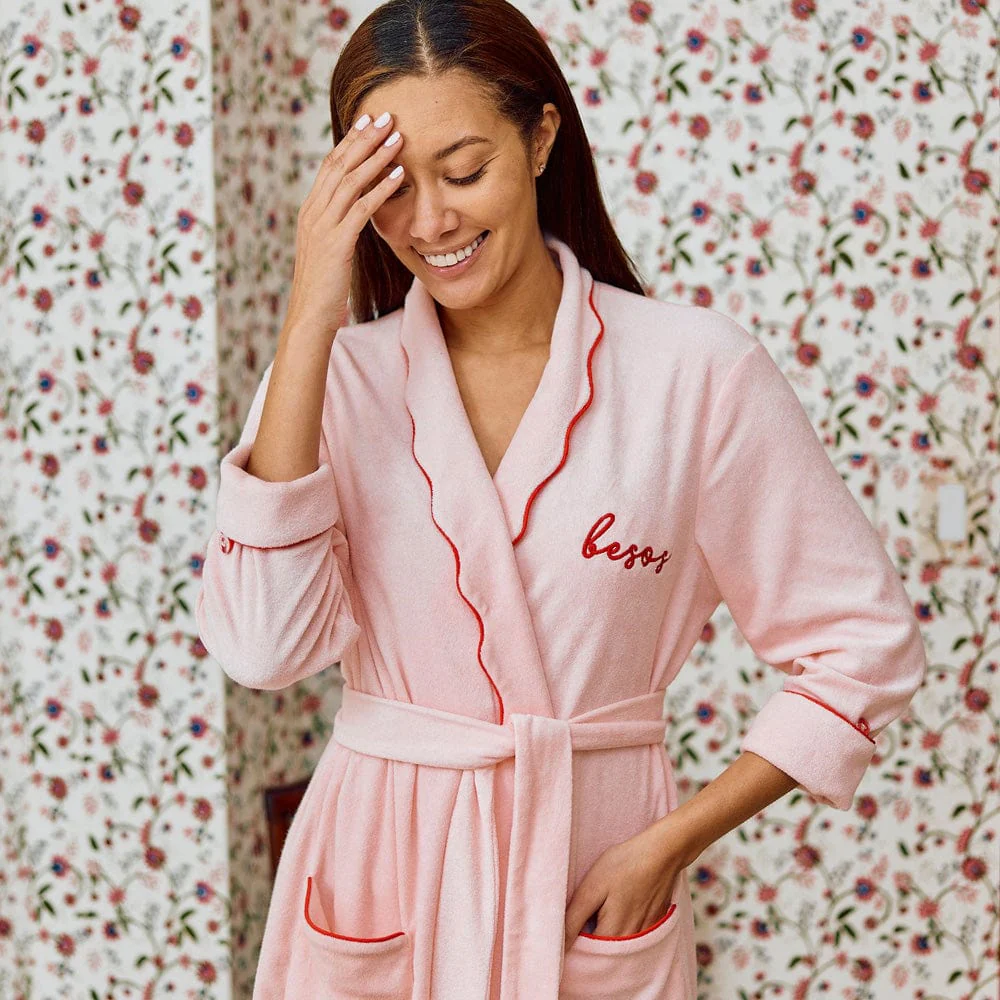 Woman wearing a pink robe with 'besos' embroidery in a floral room.
